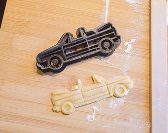 May include: A black plastic convertible car-shaped cookie cutter next to a cookie cut in the same shape. The cookie and cutter are on a wooden surface with flour.