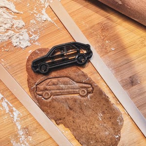 Puede incluir: Un cortador de galletas negro con forma de coche se encuentra sobre una galleta recortada, junto a un rodillo y guías de madera. La galleta está sobre una superficie de madera espolvoreada con harina.