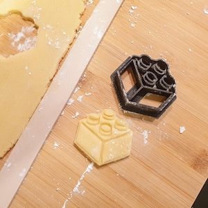 Puede incluir: Un cortador de galletas de plástico negro con forma de bloque de construcción de juguete se encuentra junto a una galleta cortada y masa enrollada. El cortador está sobre una superficie de madera, con harina esparcida.