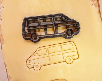May include: A black van-shaped cookie cutter sits above a van-shaped cookie cut-out on a light yellow surface. The cookie cutter is made of a dark material and has detailed windows and wheels. The cut-out is a lighter shade of yellow.