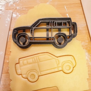 May include: A black plastic car-shaped cookie cutter sits atop a piece of dough, with an imprint of the car visible below. The cutter is on a wooden surface with a rolling pin and flour.