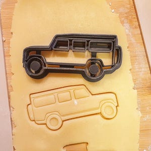 May include: A black cookie cutter in the shape of a vintage SUV sits atop a piece of dough, with an imprint of the vehicle below. The dough is on a wooden surface, with a white strip on the side.