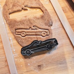 Puede incluir: Un cortador de galletas negro con forma de coche descapotable junto a un recorte en masa. La masa es de color marrón claro y el cortador es de plástico negro. La imagen está sobre una superficie de madera con utensilios de repostería.