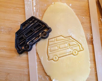 May include: A black plastic cookie cutter in the shape of a camper van sits next to a piece of dough with the van's imprint. The dough is light yellow, and the cutter is on a wooden surface. A wooden rolling pin is in the background.