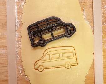 May include: A black plastic cookie cutter in the shape of a camper van sits on a sheet of dough. The dough has an imprint of a camper van. The dough is on a wooden surface.