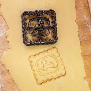 May include: A black cookie cutter with a scalloped edge sits on a sheet of dough, next to a cut-out cookie. The cookie cutter has the John Deere logo, a leaping deer inside a shield. The cut-out cookie is light brown.