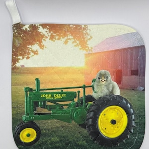 May include: A green John Deere tractor with a yellow wheel and a small white chick sitting on the seat. The tractor is in a field with a red barn in the background.