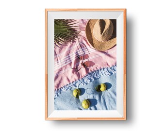 May include: A framed image featuring a beach scene. A straw hat, pink sunglasses, and a pink and blue striped beach towel are arranged on a blue and pink towel. Green fruit and a palm frond complete the scene.