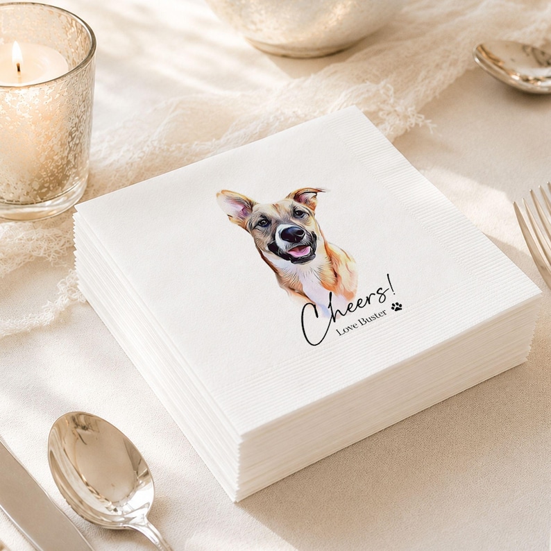 Op de afbeelding: Een stapel vierkante witte papieren servetten met een kleurrijke illustratie van een bruin-witte hond met de tong uit de bek. Onder de hond staat de tekst Cheers! Love Buster, vergezeld van een klein zwart icoontje van een pootafdruk.