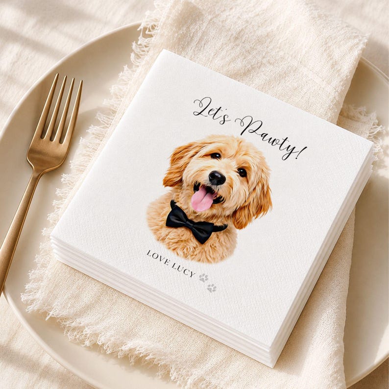 Op de afbeelding: Een wit vierkant papieren servet met een gedrukte illustratie van een goudkleurige hond met een zwarte vlinderdas. De tekst Let's Pawty! staat boven de hond en de tekst LOVE LUCY met twee kleine pootafdrukken staat eronder.