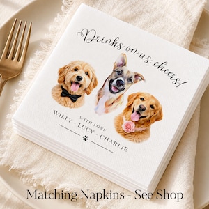 May include: A white square paper napkin featuring the printed text Drinks on us, cheers! and WITH LOVE WILLY - LUCY - CHARLIE with a paw print. The design includes three illustrated dog portraits: one wearing a black bow tie, one plain, and one with a pink flower collar.