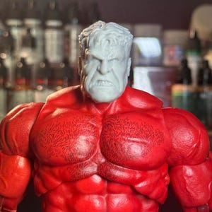 Pode incluir: Um close-up da cabeça de uma figura de ação vermelha e cinza. A figura tem uma expressão esculpida, zangada e um peito detalhado com cabelo.