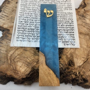 May include: A blue and brown wooden bookmark with a gold Hebrew letter "Shin" on the top. The bookmark is resting on a piece of wood.