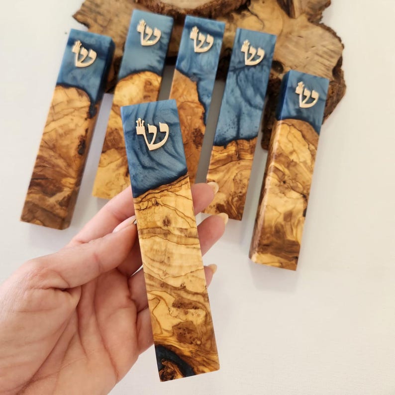 May include: A rectangular mezuzah case crafted from natural olive wood and blue resin, featuring a metallic Hebrew letter Shin on the upper portion. The wood displays a prominent grain pattern, and the blue resin has a swirling, semi-translucent appearance.