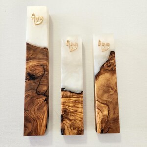 May include: Three rectangular mezuzah cases featuring a combination of natural brown wood grain and translucent white resin. Each piece is topped with a gold-colored Hebrew letter Shin, showcasing distinct organic wood patterns and smooth finishes.