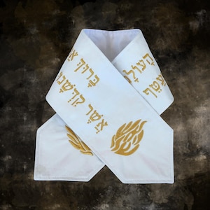 May include: White fabric item with gold Hebrew text and gold embroidered designs. The item has a folded design with text and a flame-like design on one end and a leaf design on the other end. The background is a dark, mottled brown.