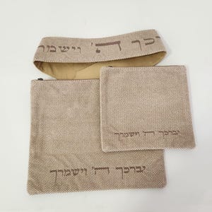 May include: Beige fabric prayer shawl bag set with Hebrew text. The set includes a rectangular bag, a smaller square bag, and a head covering. The text is in a darker shade of brown.