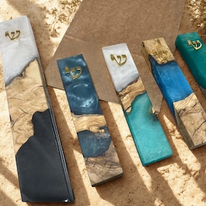 May include: Five rectangular mezuzah cases made of natural wood and colored resin. Each piece features a gold-colored Hebrew letter Shin at the top. The resin colors include white, black, deep blue, and turquoise, contrasting with the wood grain.