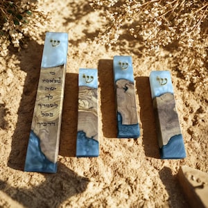 May include: Four rectangular mezuzah cases made of natural olive wood and blue resin. Each case features a gold-colored Hebrew letter Shin at the top. The tallest case displays Hebrew text engraved on the wood surface in a vertical orientation.