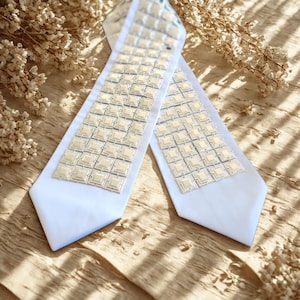May include: A white necktie featuring a central panel of interconnected, square-shaped metallic silver tiles. The tiles have a raised, pyramid-like center and a textured border, creating a geometric pattern that runs down the length of the tie.