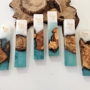 May include: Six rectangular mezuzah cases made of olive wood and teal resin with white swirls. Each case features a gold-colored Hebrew letter Shin at the top. The wood grain is visible within the resin, and the cases are arranged in a vertical row.