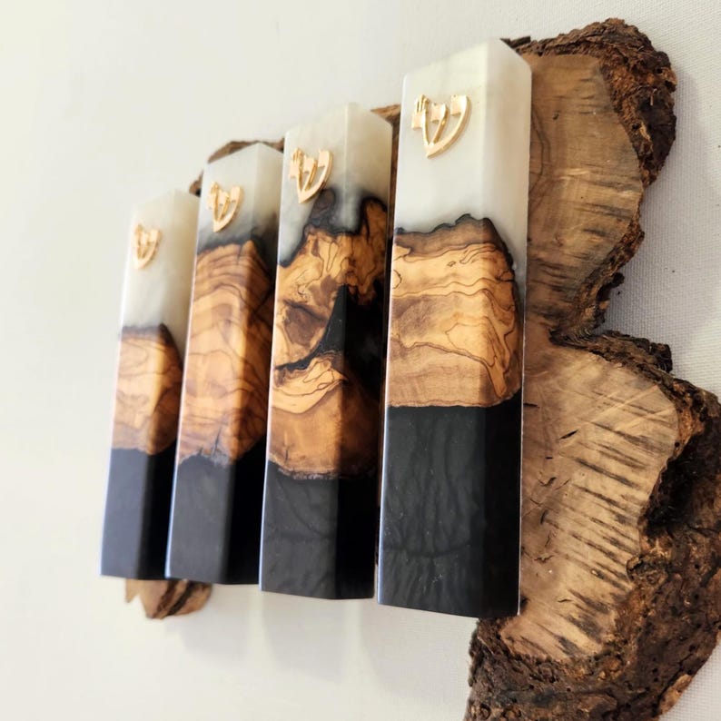 May include: Four rectangular mezuzah cases arranged vertically, each featuring a combination of light cream resin, natural olive wood with visible grain, and solid black resin at the base. Each case is topped with a small gold-colored Hebrew letter Shin.