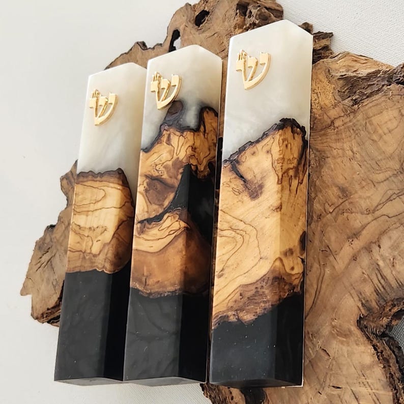 May include: Three rectangular mezuzah cases made of a combination of natural wood with visible grain, opaque white resin, and solid black resin. Each case features a gold-colored Hebrew letter Shin attached to the upper white resin section.
