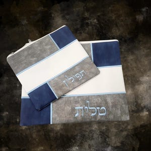 May include: Two rectangular prayer bags, one zippered, in a patchwork design of grey, white, and navy blue. The bags have Hebrew text embroidered in light blue. The background is a dark, mottled grey.