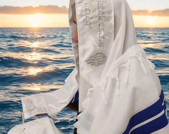 Puede incluir: Un chal de oración tallit blanco con rayas horizontales azules y bordados plateados. La atará muestra texto en hebreo y un motivo de llama. La prenda incluye flecos anudados en las esquinas y una confección de tela texturizada.