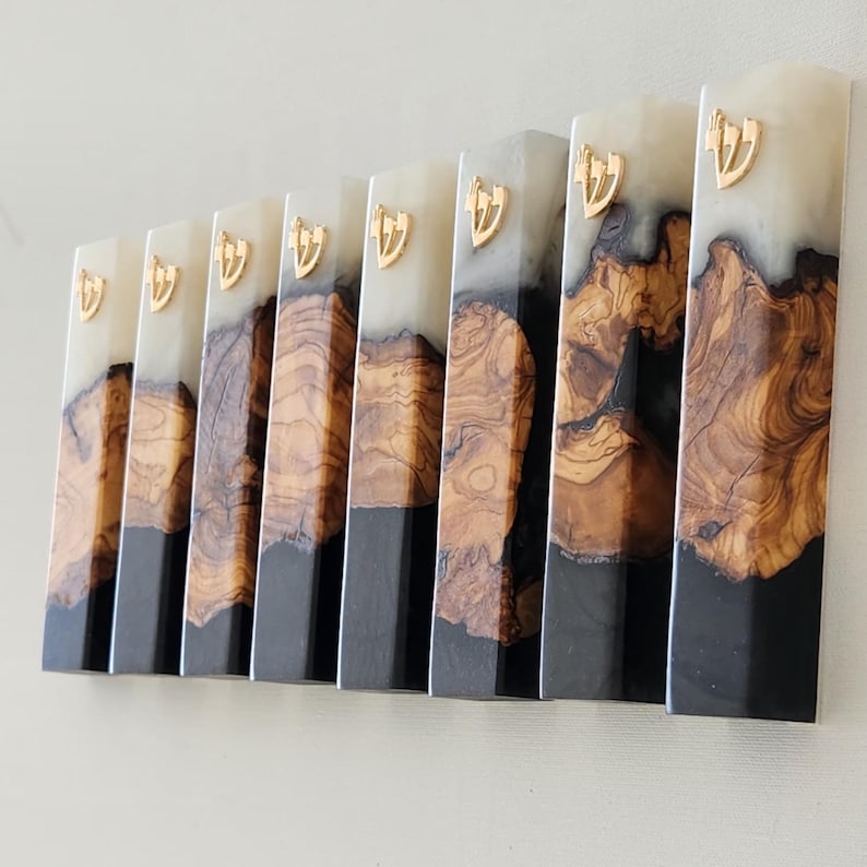 May include: A collection of eight rectangular mezuzah cases arranged in a row. Each case features a combination of natural wood grain at the center, a solid black base, and a translucent white top section, accented with a gold-colored Hebrew letter Shin.