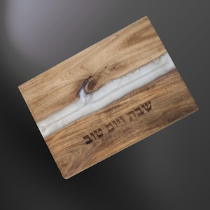 Puede incluir: Tabla de cortar de madera con un diseño de río de resina blanca. La tabla rectangular tiene un patrón de vetas de madera natural. El texto "Shabbat Shalom" está grabado en la madera. Ideal para servir alimentos.