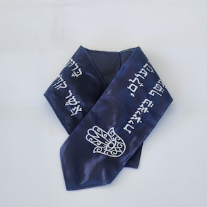 May include: A navy blue scarf with white Hebrew text and a Hamsa hand design. The scarf is made of a shiny fabric and is folded in a V-shape.