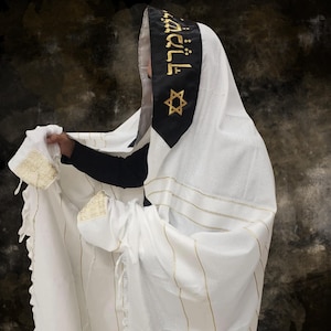 May include: White prayer shawl with gold stripes and trim. The shawl has a black collar with gold Hebrew text and a gold Star of David. The person is wearing a black shirt under the shawl.