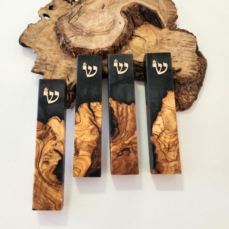 May include: Four rectangular mezuzah cases made from a combination of natural olive wood with its distinct grain patterns and smooth black resin. Each case features a gold-colored Hebrew letter Shin attached to the upper portion of the black resin section.