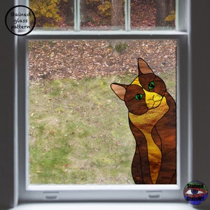 May include: A stained glass window design featuring a brown and yellow cat with green eyes, looking out of a window. The cat is positioned in the lower right corner of the window.
