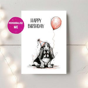 May include: A white birthday card featuring a Basset Hound illustration wearing a party hat and holding a red balloon. The words "HAPPY BIRTHDAY" are printed above the dog. A pink circle with the words "PERSONALISE ME" is in the upper left corner.