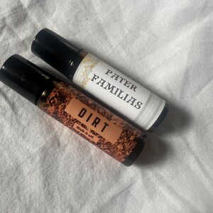 May include: Two roll-on bottles with black caps. One label reads "PATER FAMILIAS" in black text on a white background. The other label reads "DIRT" in white text on a brown background, with "Walk it off." below.