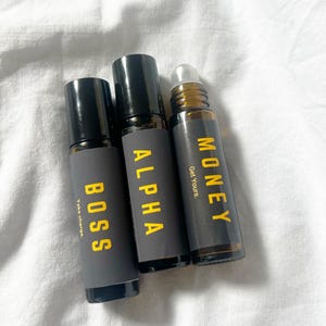 May include: Three essential oil roller bottles with black caps. The bottles are labelled "BOSS", "ALPHA", and "MONEY" in yellow text. The "BOSS" bottle says "Take charge." The "MONEY" bottle says "Get Yours."
