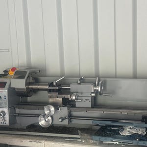 Peut inclure: Un tour à métaux gris avec divers cadrans, leviers et un affichage numérique. La machine est posée sur une table avec une boîte à outils et des outils. Le tour est conçu pour façonner le métal et d'autres matériaux. La longueur de la machine est d'environ 100 cm.