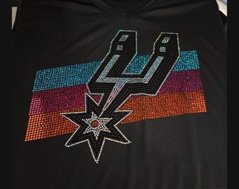 May include: Black V-neck t-shirt featuring a rhinestone design of the San Antonio Spurs logo. The logo includes a silver spur, a lightning bolt, and a star, set against a backdrop of horizontal stripes in teal, purple, and orange.