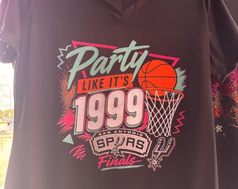 May include: A black V-neck t-shirt featuring a graphic print with the text "Party LIKE IT'S 1999" in teal and orange. The design includes a basketball, a hoop, and the San Antonio Spurs Finals logo in white, pink, and black colors.