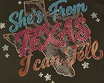 May include: Black t-shirt with a rhinestone design. The design features the text "She's From Texas I Can Tell" in blue, pink, and orange rhinestones, with a rhinestone outline of the state of Texas.