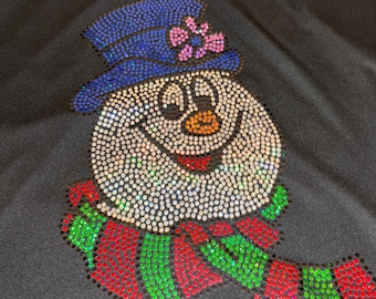 May include: Black V-neck shirt with a rhinestone snowman design. The snowman has a blue hat with a pink bow, an orange carrot nose, and a red, green, and white striped scarf. The snowman's face and body are covered in rhinestones.