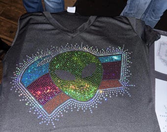 May include: A black V-neck t-shirt featuring a central alien head graphic composed of shimmering green, blue, and purple rhinestones. The design is surrounded by a radiating border of clear crystals, creating a sparkling effect on the dark fabric.