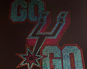May include: A black t-shirt featuring the text GO GO in a block font, embellished with iridescent rhinestones in shades of blue, pink, and orange. A stylized spur logo is positioned between the words, also decorated with matching rhinestones.
