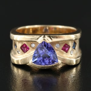 Könnte beinhalten: Ein goldener Ring mit einem großen, dreieckigen, lila und blauen Edelstein, umgeben von kleineren roten und blauen Edelsteinen und Diamanten.