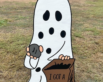 May include: A white wooden ghost cutout with black eyes and mouth, holding a gray rock and a brown bag that says "I GOT A ROCK" in black letters. The cutout is on a grassy lawn.