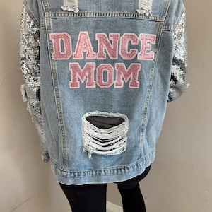 Puede incluir: Una chaqueta vaquera azul claro con mangas de lentejuelas plateadas y un parche rosa "DANCE MOM" en la espalda.