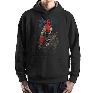 Op de afbeelding: Zwart hoodie met een grafische print van een roodhuidige figuur met een groot hoofd en hoorns, omringd door andere figuren en objecten. Het figuur draagt een bruine jas en houdt een pistool vast.