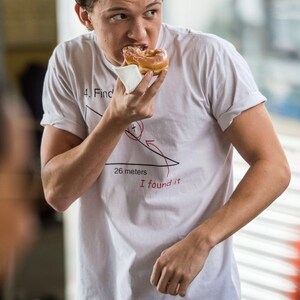 May include: A young man wearing a white t-shirt with a red drawing of a triangle and the text "4. Find 26 meters I found it" is eating a glazed donut.